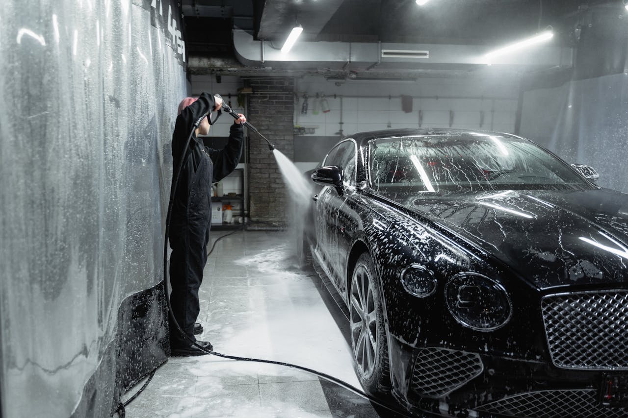 A person in a garage power washing a black car covered in soap and water spray.